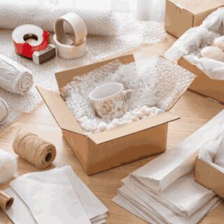 Open cardboard box filled with packing peanuts and bubble wrap, holding a carefully wrapped porcelain cup, surrounded by packing pape