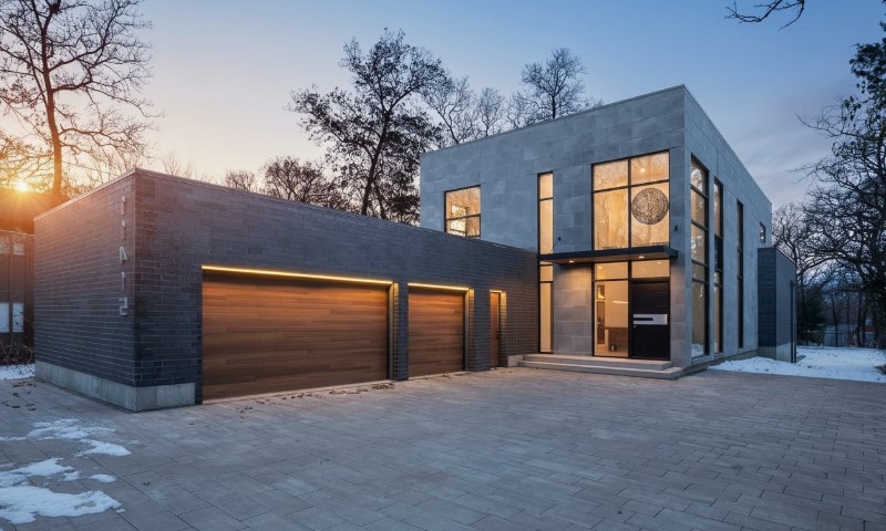 Modern house with a sleek design at dusk. Features include large windows, illuminated double garage, and snow-dusted surroundings
